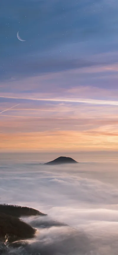 Clouds lap like water around one dark hilltop at dusk, a thin moon high in the left corner; most of the frame is soft, even gradient.