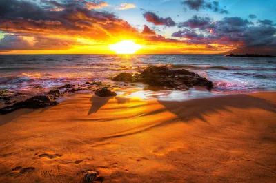 Low light rakes across rippled golden sand toward a rocky outcrop and a flaring sun, spray hanging over the waves on the left.