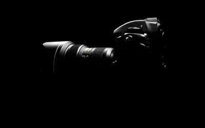 The outline of a DSLR and its long zoom lens emerges from darkness in monochrome, everything right of centre fading to solid black.