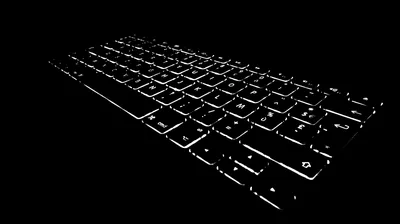 A laptop keyboard glows from below at a low angle, the keys the only thing visible in an otherwise black frame.