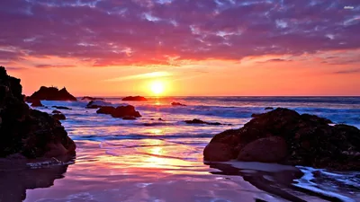 Seaweed-dark rocks frame a channel of wet sand that mirrors the pink and purple clouds, the sun sitting small and bright on the centre horizon.