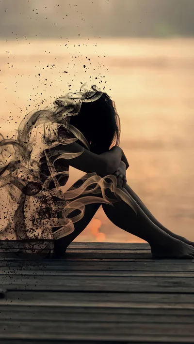 Seated on dock boards with knees drawn up, a woman's outline breaks apart into dark particles streaming left, the warm blurred water behind her out of focus.