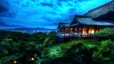 Lantern light spills from a wooden temple perched over dark forest at blue hour, city lights glowing far left; the subject sits right of centre in this landscape frame.