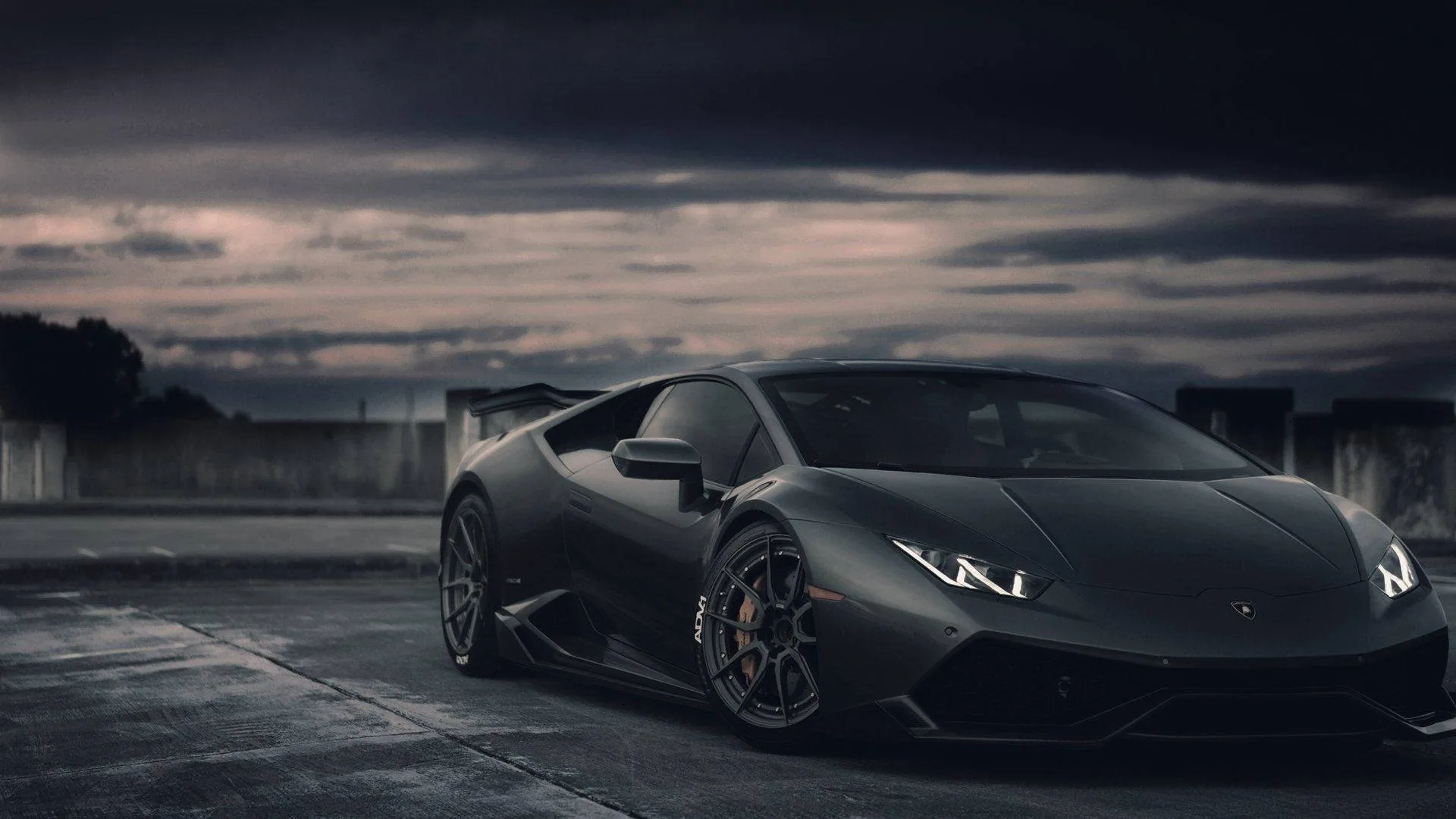 Charcoal Lamborghini parked side-on under a bruised evening sky, brake discs and hex wheels sharp while the background fades to grey.