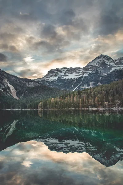 Broken evening clouds and a snow-capped ridge mirror sharply in dark green water, the symmetrical reflection filling the lower half of this portrait frame.
