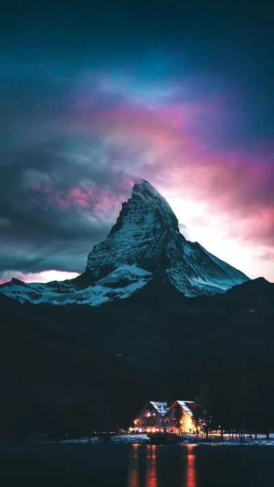 A snow-dusted rock pyramid rises into pink and grey dusk clouds, a small lit chalet at the shoreline spilling gold reflections across the bottom edge.