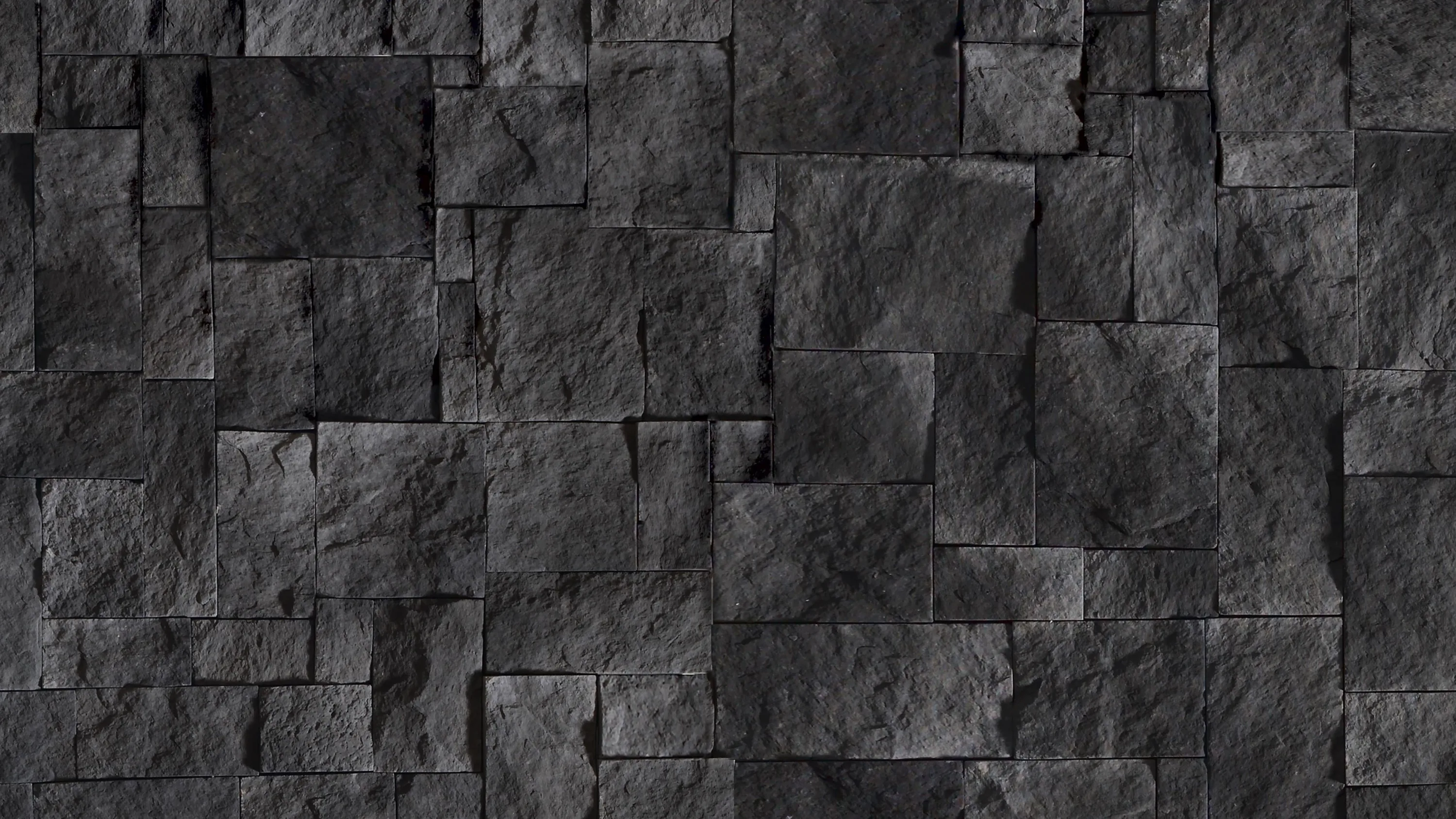 Rough-cut charcoal stone blocks interlock in staggered courses, side light raking across so every chipped edge throws its own shadow.