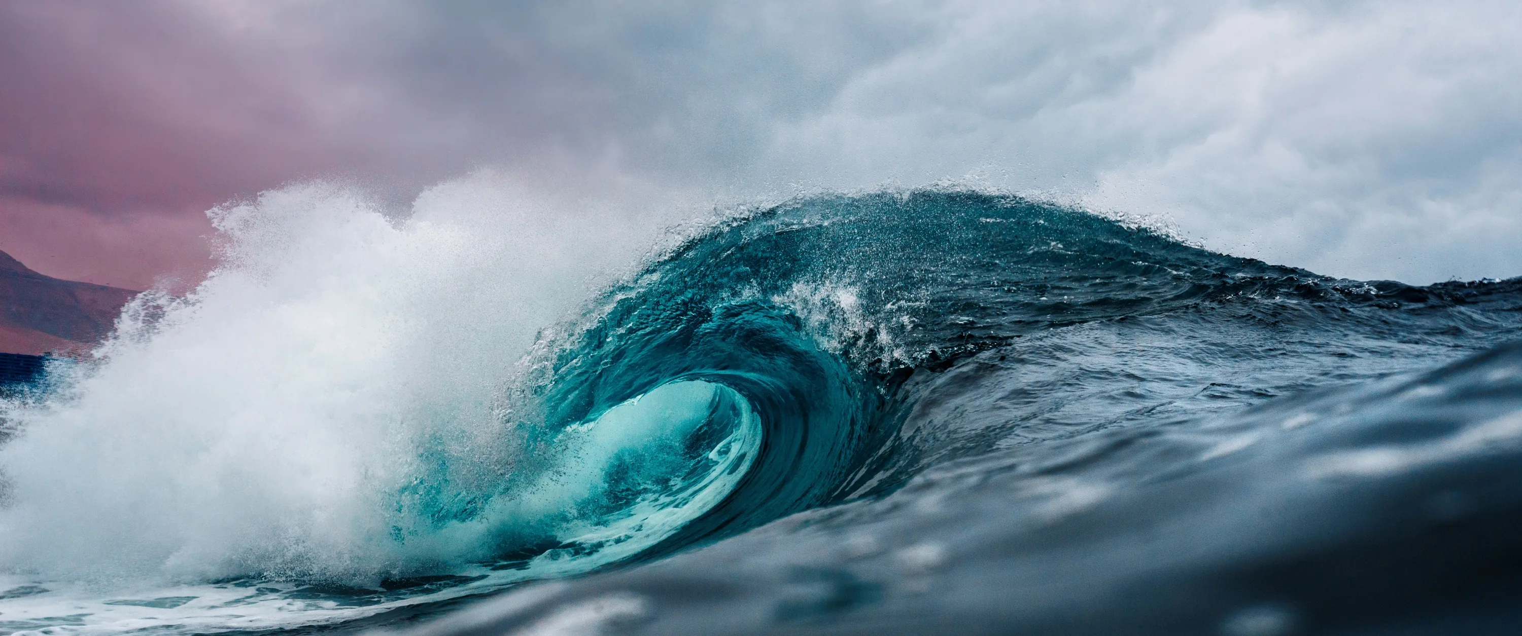 A glassy turquoise barrel curls over against a grey-pink storm sky, spray blowing back off the crest in an ultrawide crop.