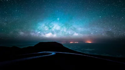 The Milky Way glows green over a dark volcanic summit, a road curving through the bottom of the frame toward distant orange cloud.