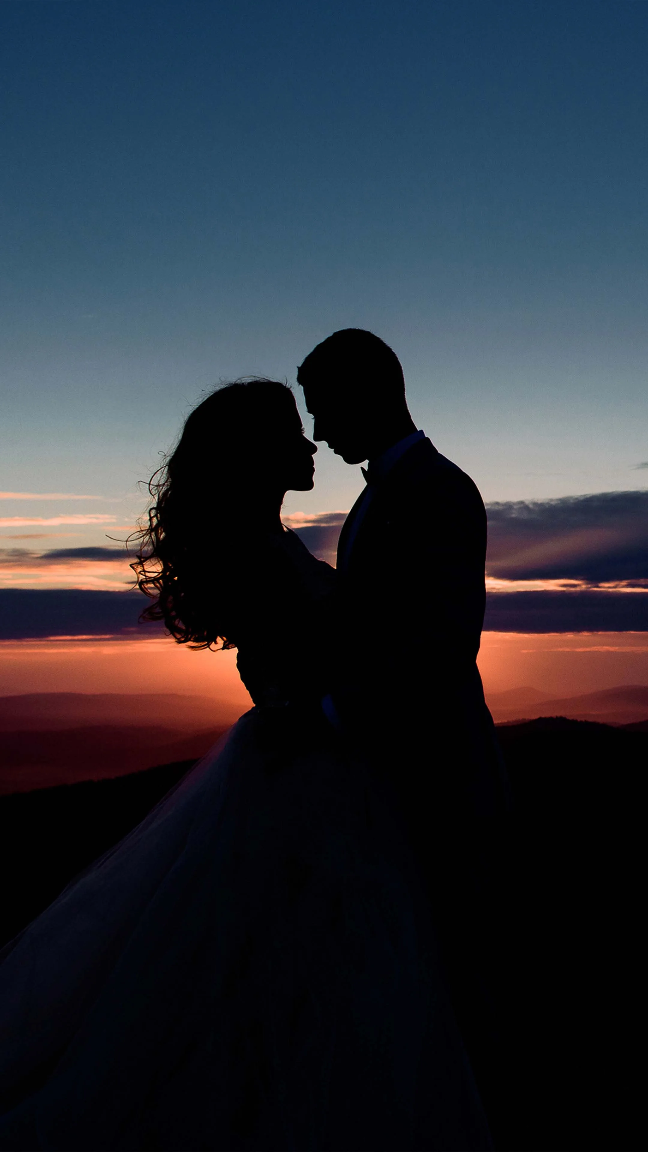 A couple stand forehead to forehead on a hilltop, her full skirt and his suit reduced to flat black against a band of orange cloud under deep blue sky.
