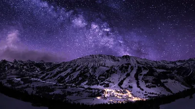 The galactic band leans over a broad snowy massif, a village glittering at its foot — most of the brightness stays in the bottom centre.