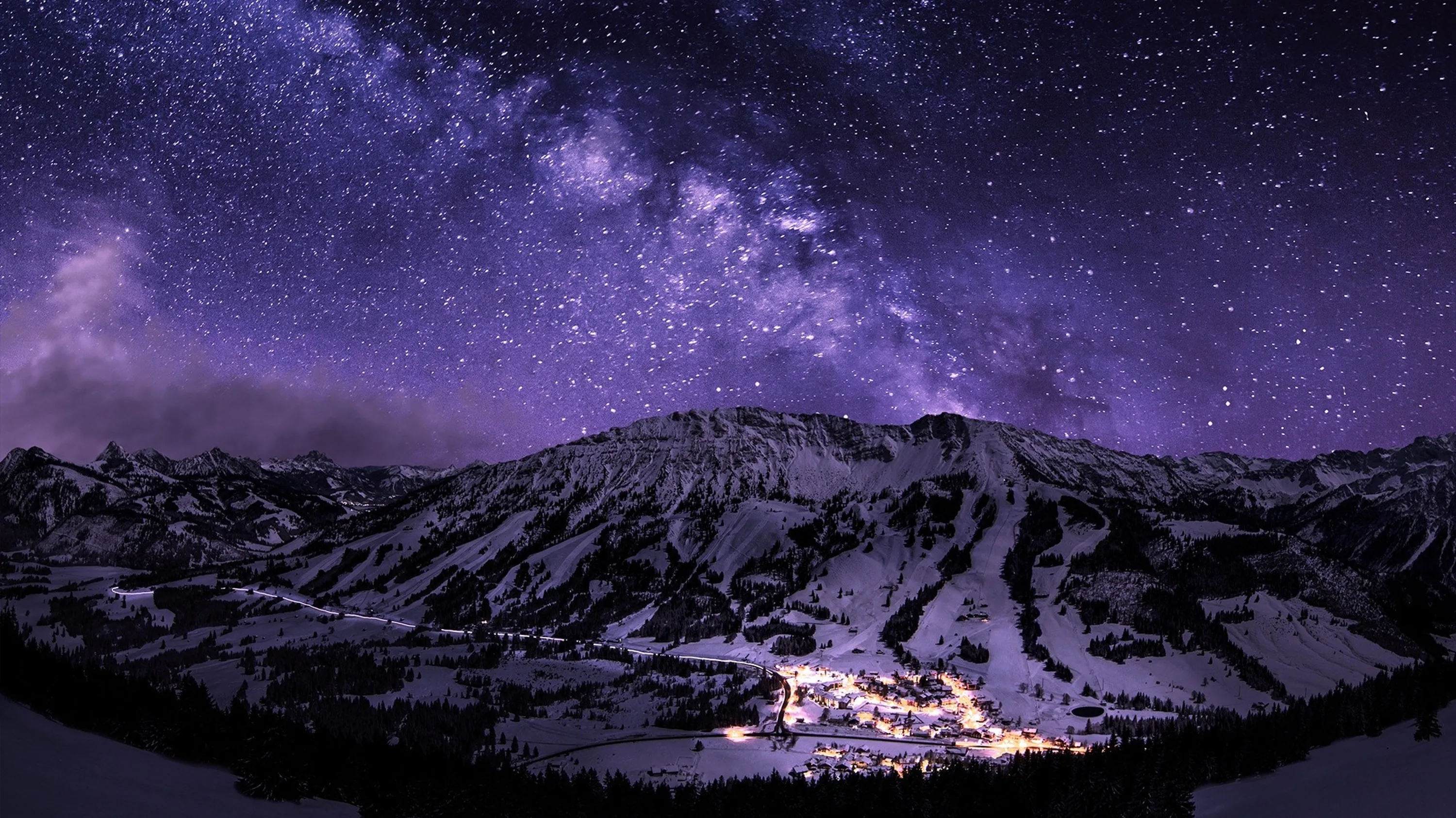 The galactic band leans over a broad snowy massif, a village glittering at its foot — most of the brightness stays in the bottom centre.