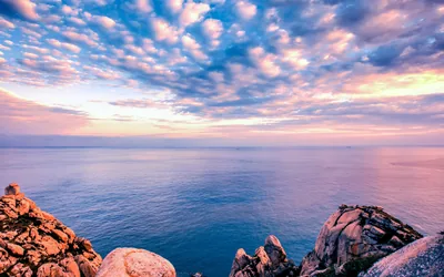 Rows of small pink clouds scatter over a calm blue sea at sunset, with rocky outcrops anchoring the lower right corner.