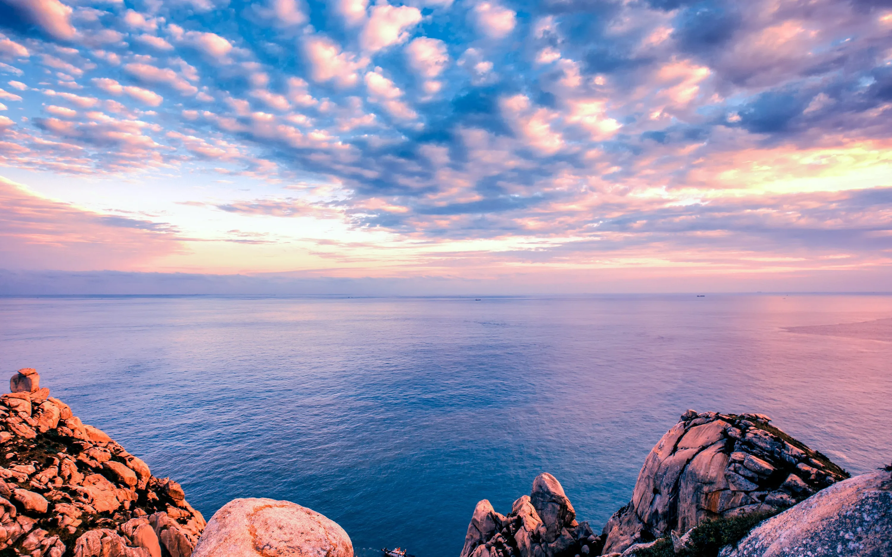 Rows of small pink clouds scatter over a calm blue sea at sunset, with rocky outcrops anchoring the lower right corner.