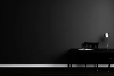 A black wall fills most of the frame with a desk, chair and small chrome lamp pushed to the right, an open book under the light.