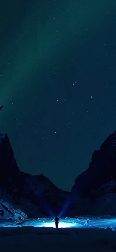 A tiny figure shines a beam into the dark between two mountain walls, cold blue light pooling at the base while faint aurora tints the sky.