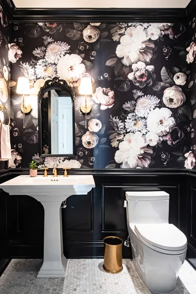 Oversized blush and white blooms on a near-black powder room wall, lit warm by sconces flanking the mirror above a pedestal sink.