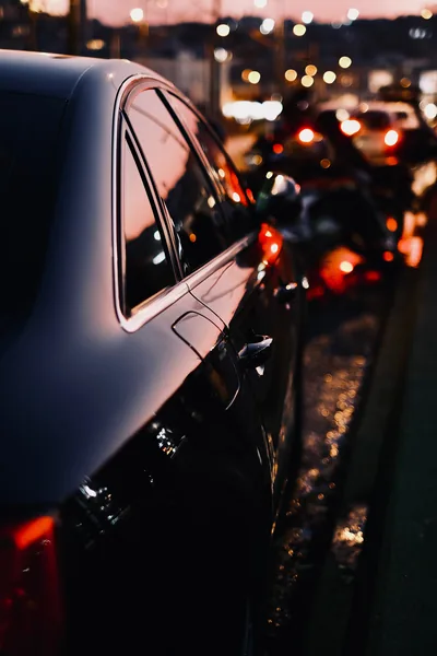 Vertical shot along the flank of a black saloon in queued traffic, a long string of blurred red brake lights and city glow filling the road ahead.