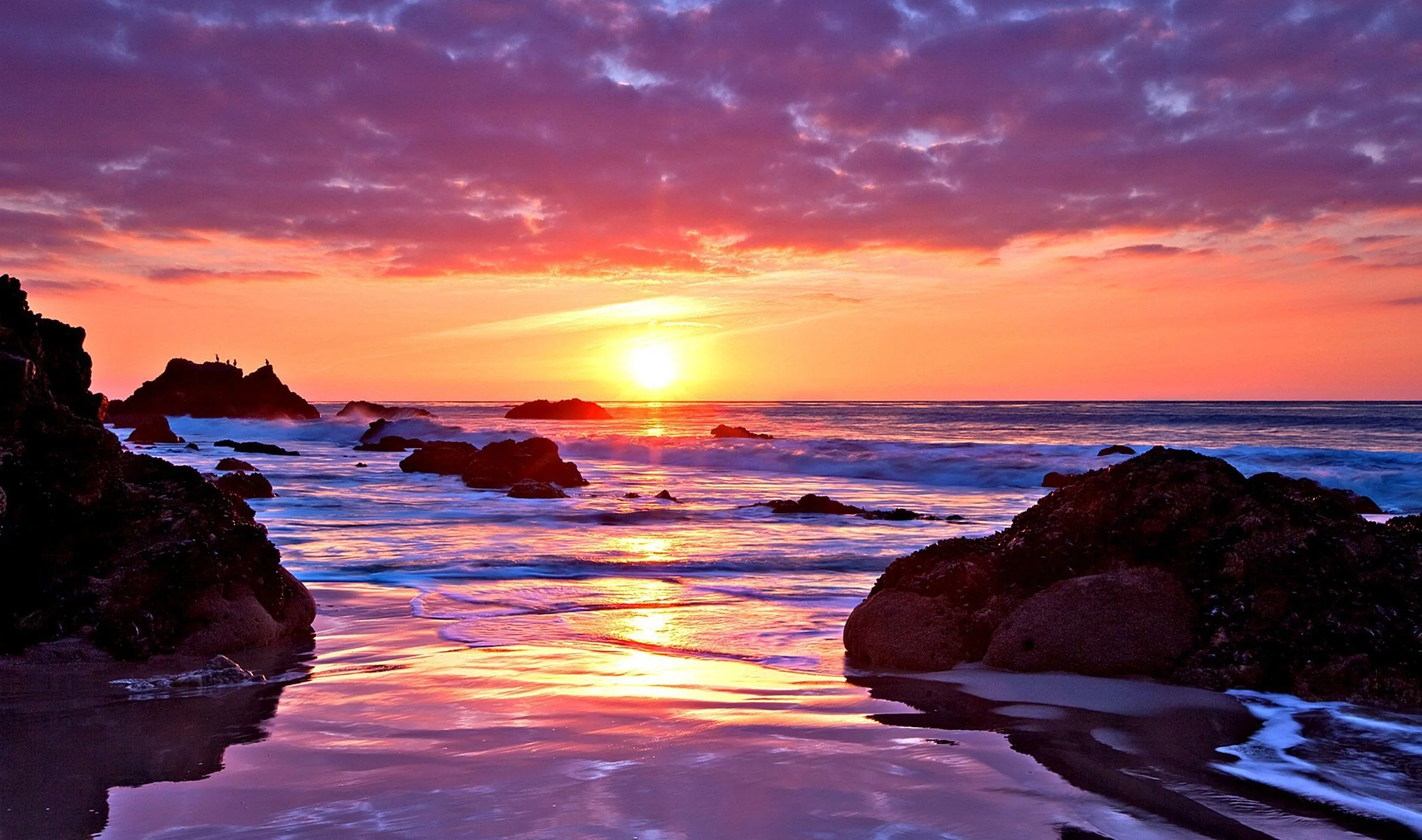 The sinking sun spills magenta and violet across wet sand and scattered rocks, its reflection running straight down the middle of the frame.