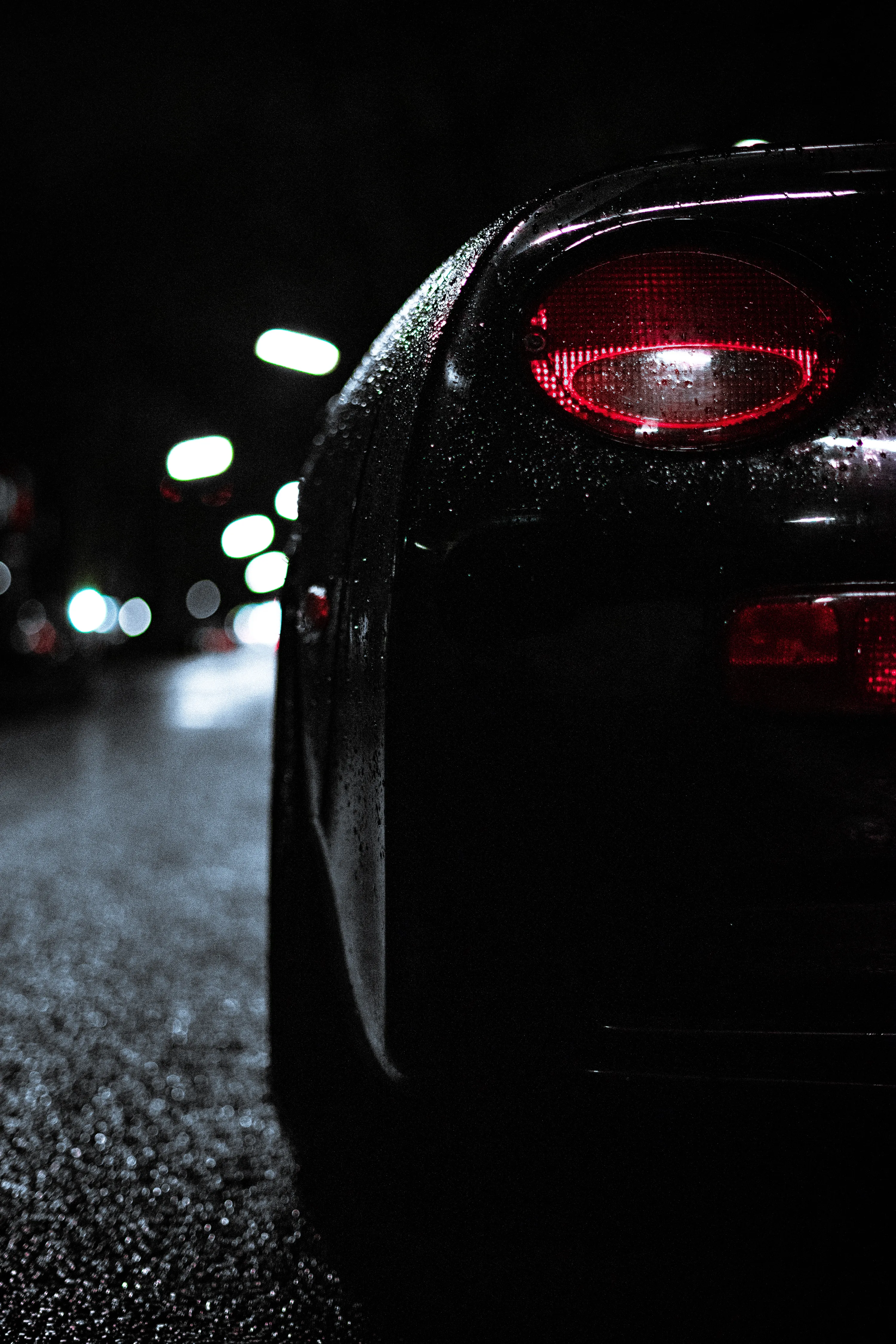 Rain beads across the rear quarter of a dark coupe, its red tail lamp burning against out-of-focus street bokeh down the road.