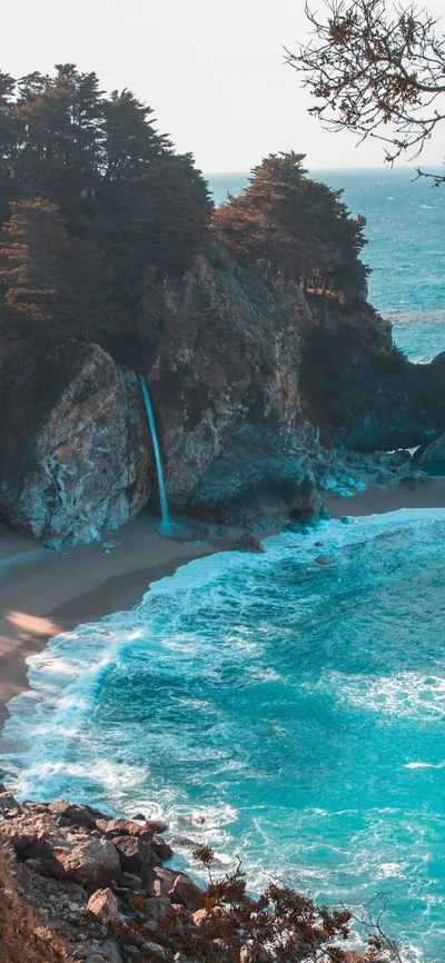 A slim waterfall drops from the cliff onto the beach left of centre, turquoise surf churning through the lower half while cliffside trees darken the top corners.