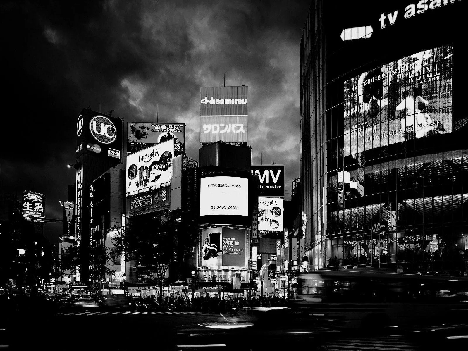 Lit billboards climb the dark towers around a wide monochrome intersection, storm clouds pressing down from above while traffic light trails streak the bottom edge.