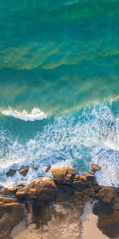 Straight-down view of surf foaming over brown rocks, the green water sliding to deeper teal along the top of the frame.