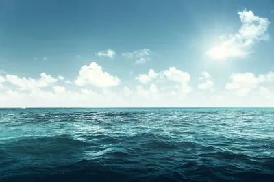Deep teal swell rolls toward the camera under scattered clouds, the sun flaring in the top right corner of the frame.