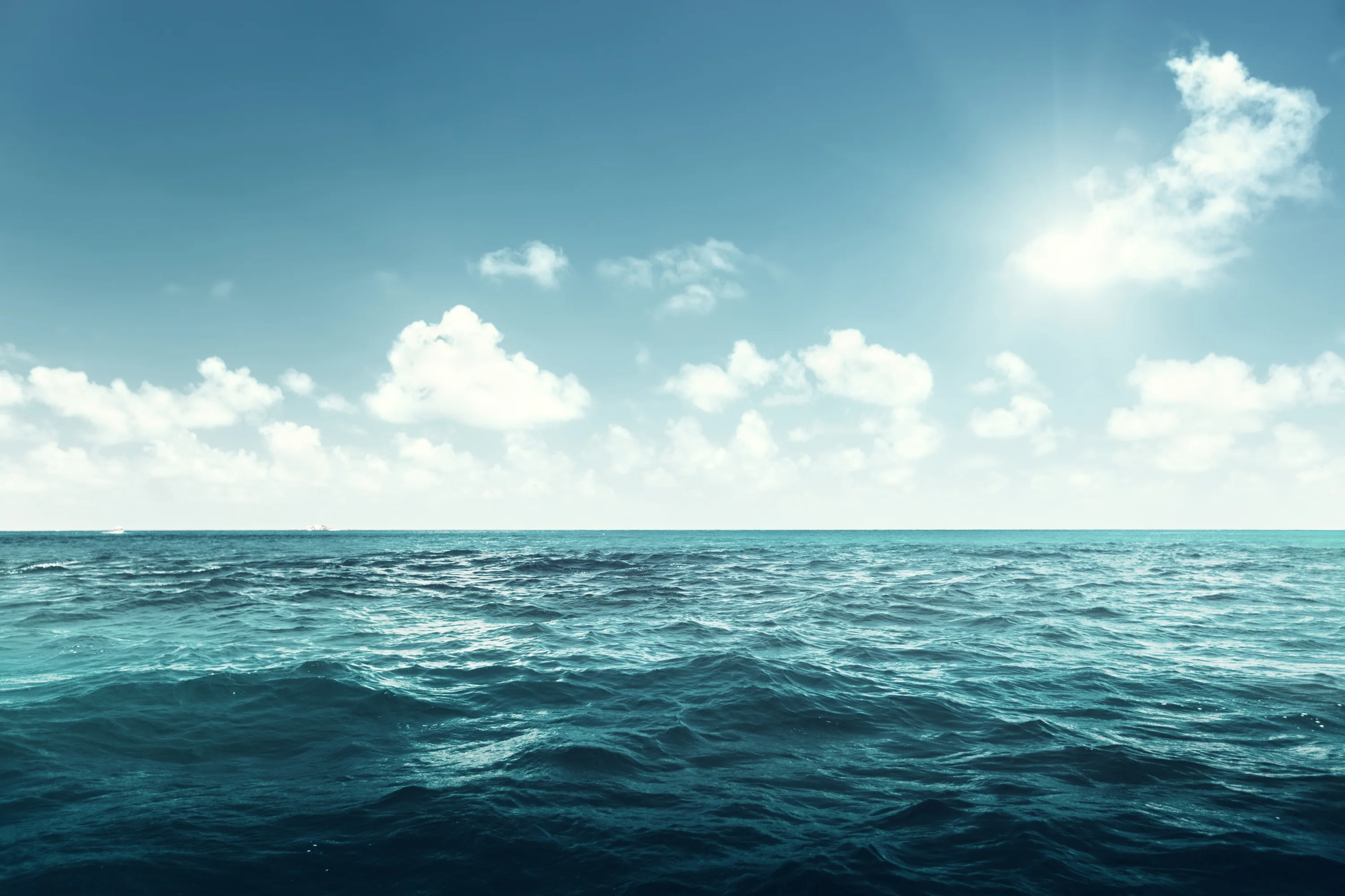 Deep teal swell rolls toward the camera under scattered clouds, the sun flaring in the top right corner of the frame.