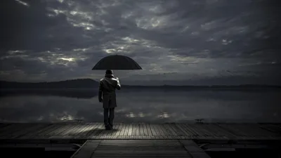 Moonlight breaks through heavy cloud onto the rim of an umbrella held on a lakeside pier, the water on the left flattening to a dark mirror.