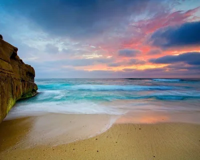 An orange sandstone bluff anchors the left edge while soft-blurred waves and pink clouds fill the rest, the open sand below suiting a dock or icons.