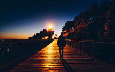 Warm light floods down the centre of this long pier as the sun drops behind the treeline, the walking figure placed dead centre so side crops survive.