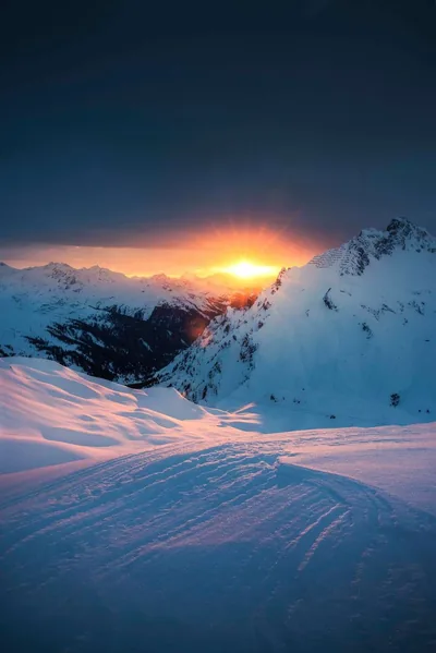 The setting sun burns orange in a notch between dark peaks, casting pink light across rippled snow while the shadowed sky stays clear for a clock.