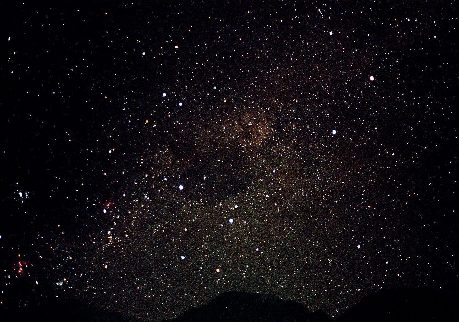 A mountain ridge cuts a black line along the bottom while the Milky Way rises overhead, faint red nebulae glowing near the left edge.