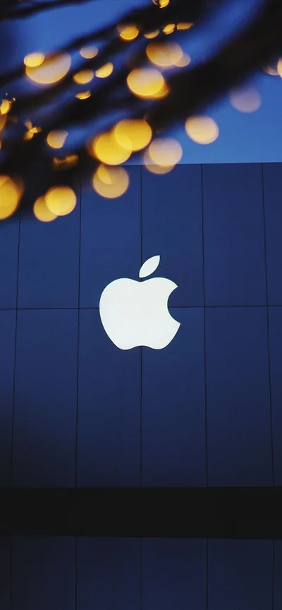 The Apple logo glows white on a panelled blue wall, with warm bokeh from string lights blurred across the top of the frame.