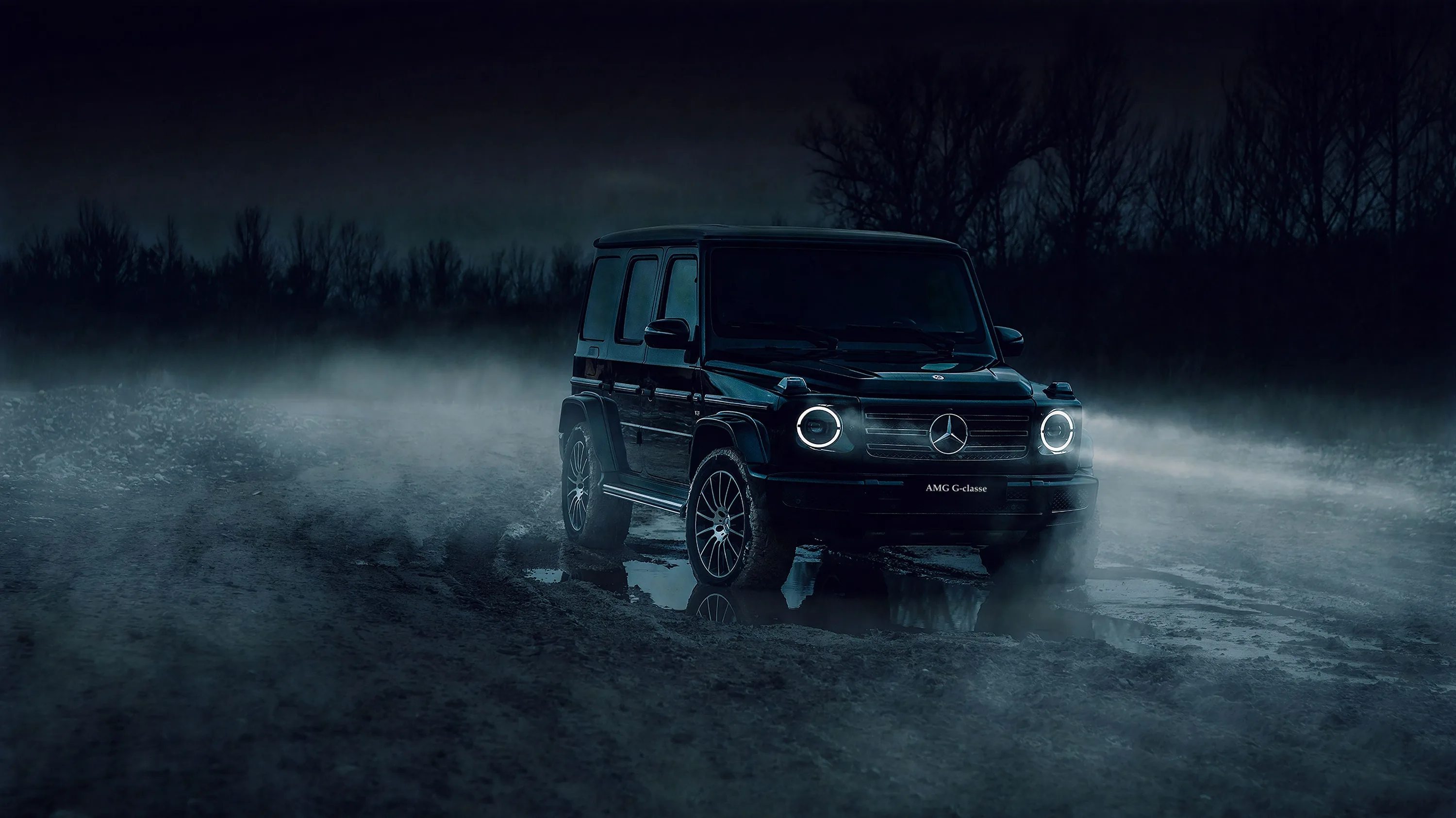 The boxy G-Wagon sits on wet ground with mist rolling past its wheels, only the round headlights and a treeline showing against the blue-black night.