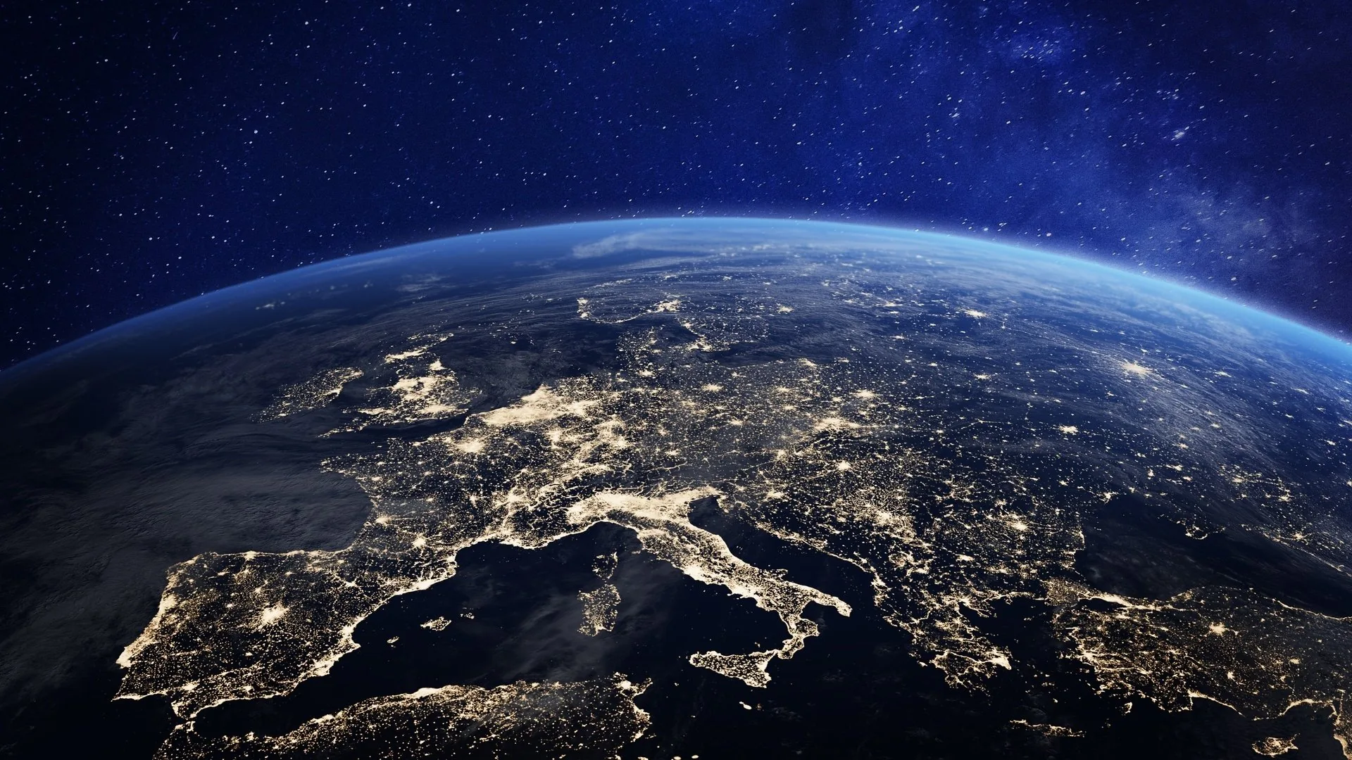 Italy, the Alps and the Mediterranean glow with city lights under thin cloud, the curve of the atmosphere fading into a starry upper edge.