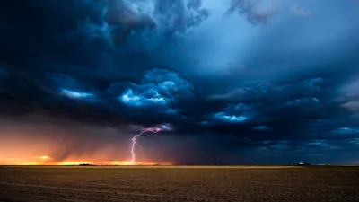 A single bolt drops from a blue-black storm cell onto flat farmland, warm town lights burning low on the left horizon.