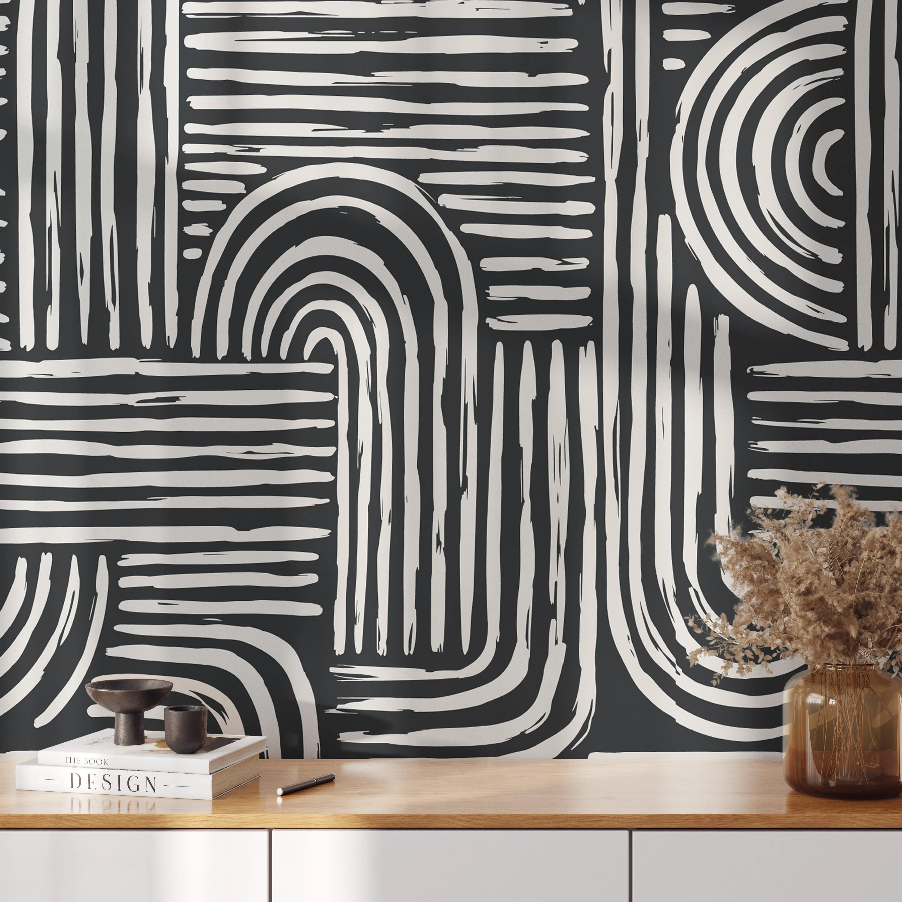 Broad brushed arcs and stripes in off-white cover a charcoal wall behind a pale wooden sideboard holding dried grasses and stacked books.