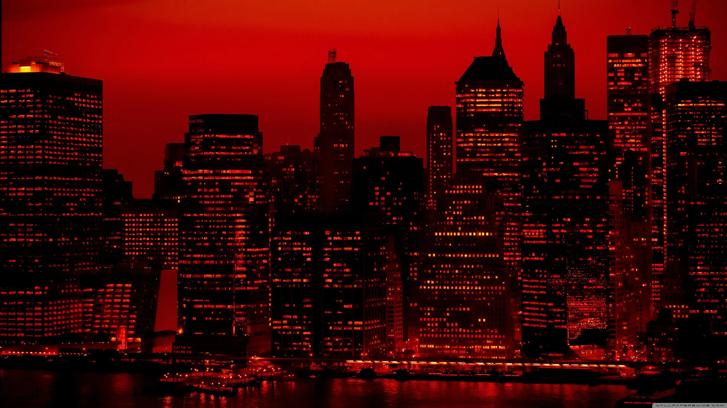Skyscrapers crowd the waterfront under a solid red sky, their windows the only pinpricks of light across an otherwise black mass.