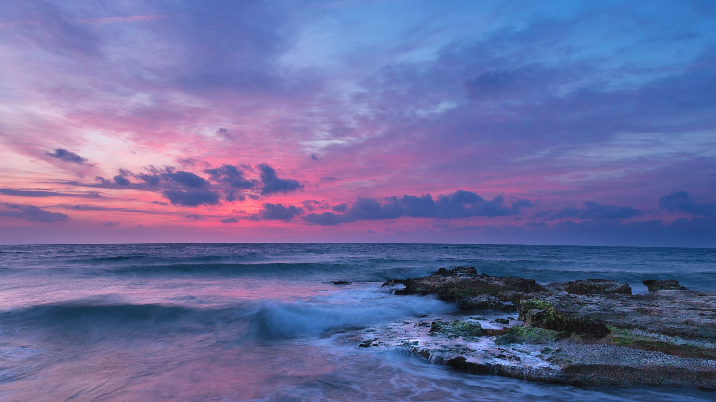 Long-exposure water smooths to mist around dark seaweed-covered rocks under a pink and violet sky, the haze softening everything but the stones.