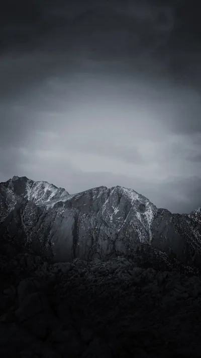 A jagged ridge holds patches of snow beneath heavy grey cloud, the dark foreground rocks falling into shadow at the bottom of the frame.