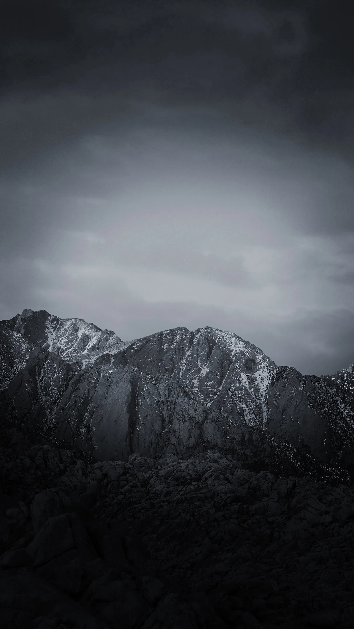 A jagged ridge holds patches of snow beneath heavy grey cloud, the dark foreground rocks falling into shadow at the bottom of the frame.