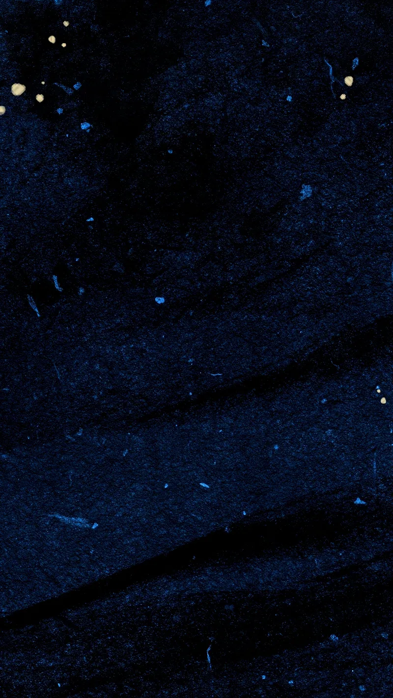 Close-up of dark indigo fabric with pale flecks and a few gold specks in the top corner, the weave creasing softly down the right.