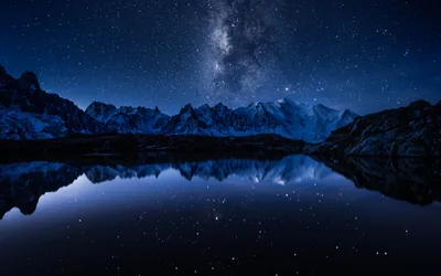 A jagged night-blue range and the Milky Way reflect in still water, the smooth dark lake across the bottom leaving room for a dock.
