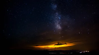 A vertical column of Milky Way dust stands centre-right while amber sunset clouds and a string of distant lights hold the horizon below.