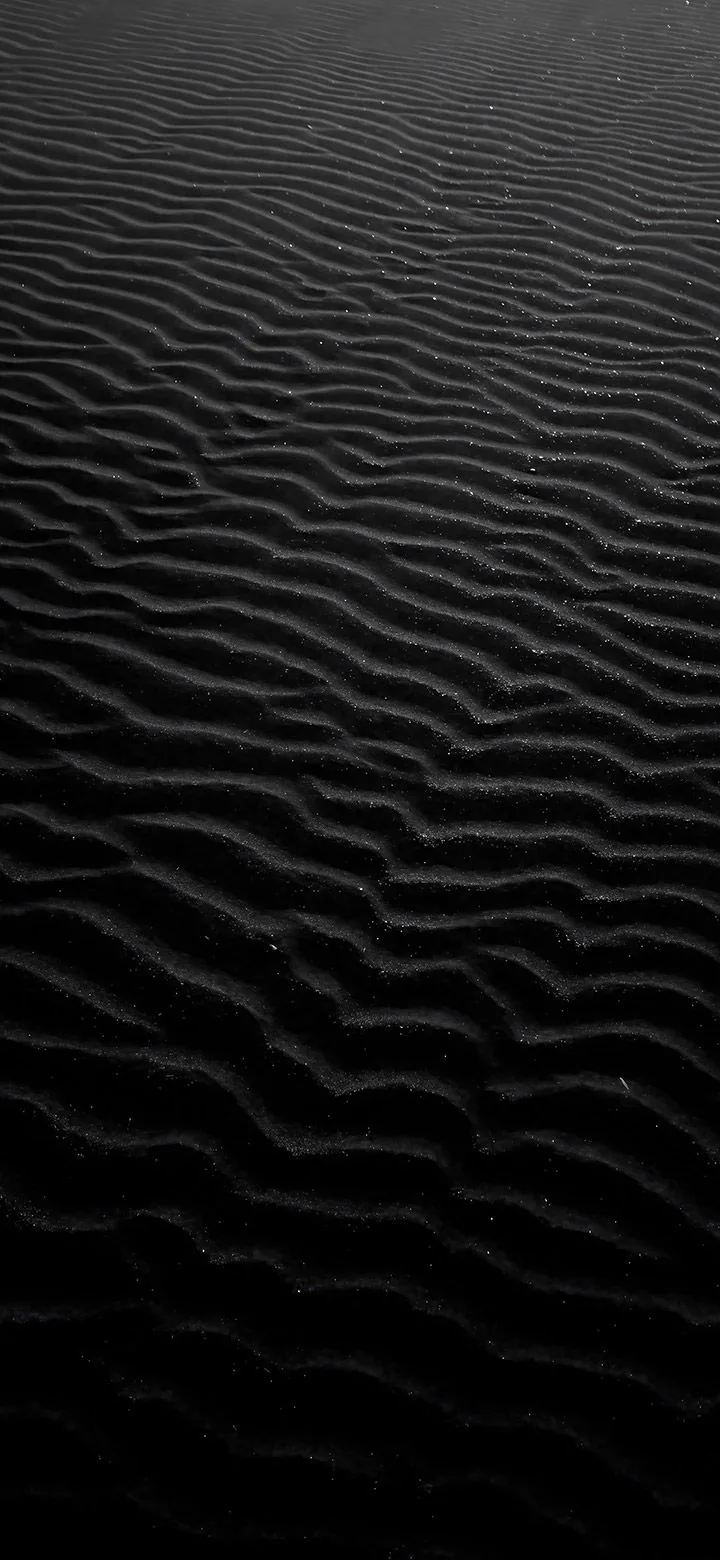 Parallel wind ripples run across dark volcanic sand toward a horizon that dissolves into black, lit from a low raking angle.