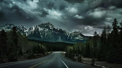 An empty highway runs dead centre toward a snow-streaked peak, storm cloud churning overhead and the pine walls on either side falling into black.