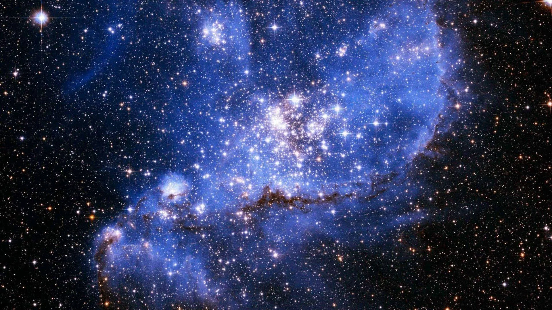 A dense cluster of newborn stars burns white at the heart of this blue nebula, surrounded by dark dust ridges and a sky packed with stars.