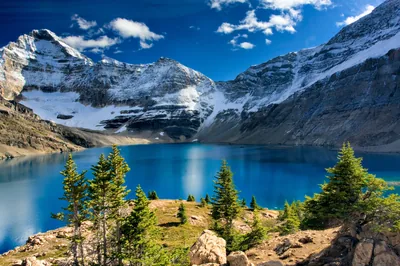 An intensely blue tarn sits in a rock cirque below snow-banded cliffs, small firs on the near slope giving the scene its scale.